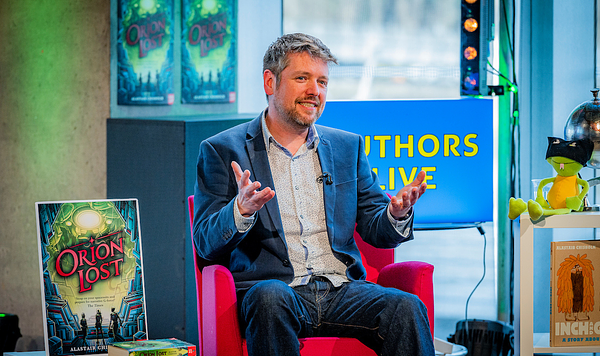 Alastair (male, brown hair, blue jacket, jeans) on the set of BBC Authors Live. Alastair is holding his hands forward as if weighing up a situation.