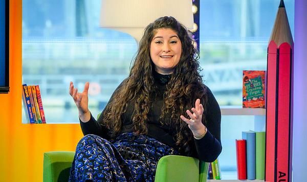 Nadine (female, long black hair, blue dress) on the set of BBC Authors Live. Nadine is holding her hands forward as if weighing up a situation.